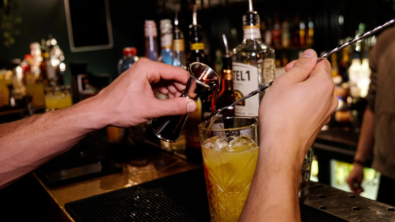 Hands stirring a tall yellow cocktail with a bar spoon in front of a stocked back bar