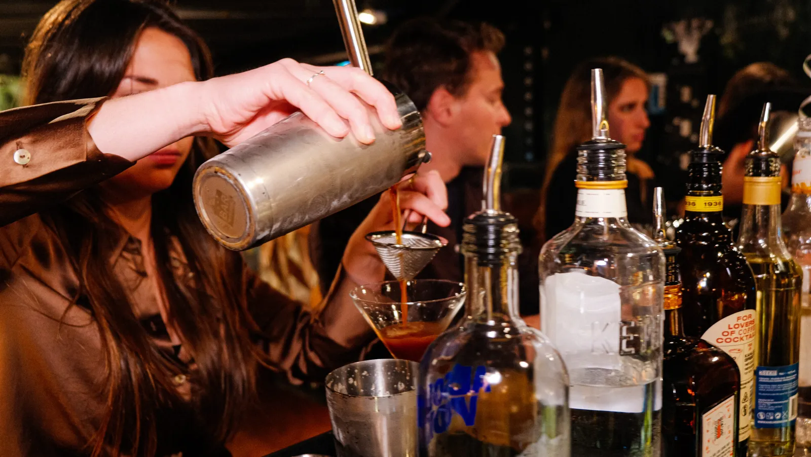 Guest straining a shaken cocktail into a glass, with bottles of spirits lined up on the bar