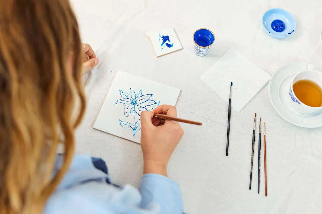 Ospite che dipinge un fiore blu su una piastrella bianca, con pennelli e una tazza di tè accanto
