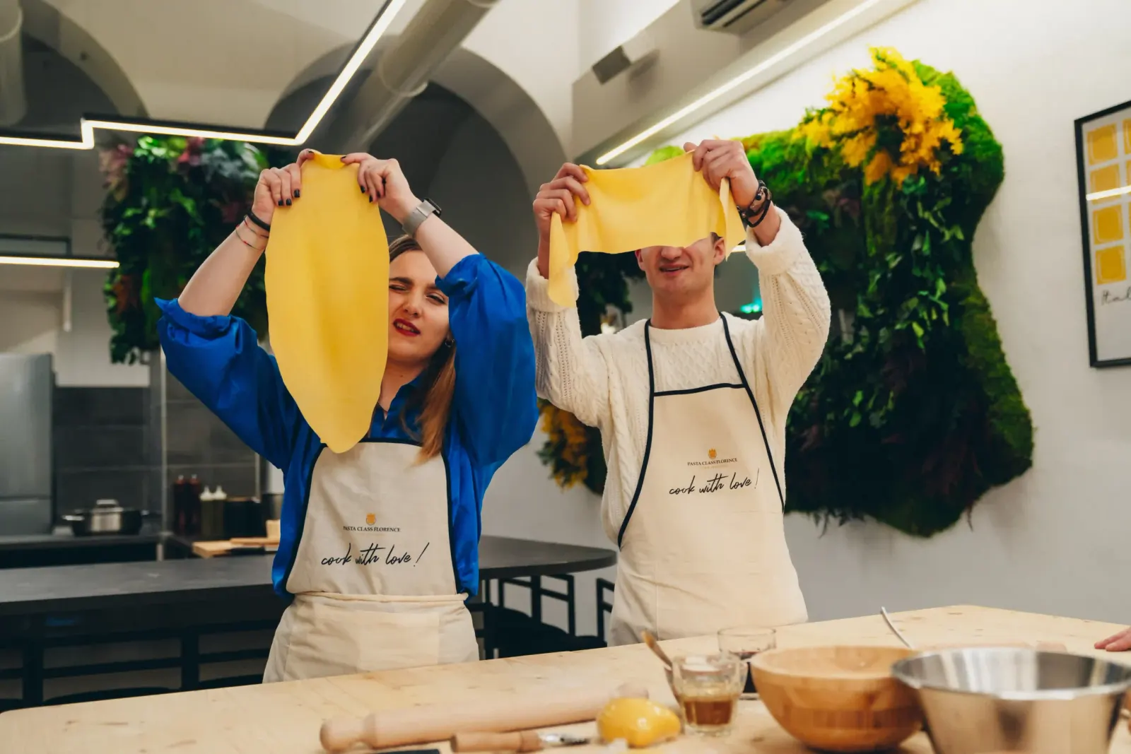 Two guests holding up wide sheets of fresh pasta dough in a bright cooking studio