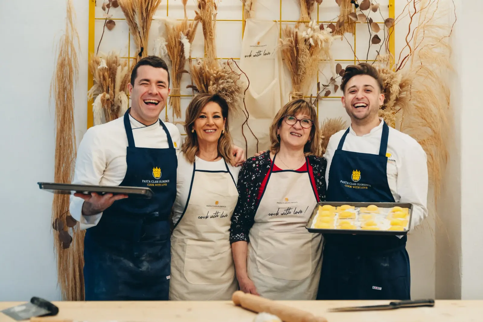 Four smiling instructors in navy aprons posing with trays of fresh pasta in front of dried flowers