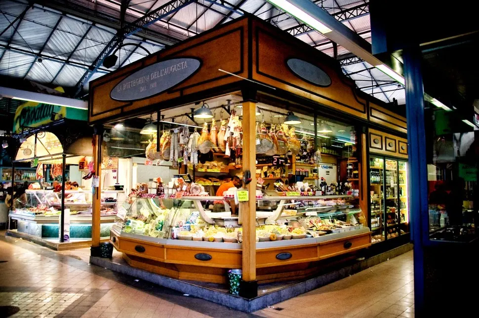 Wooden food stall under a glass-roofed market hall displaying cured meats, cheeses and prepared dishes