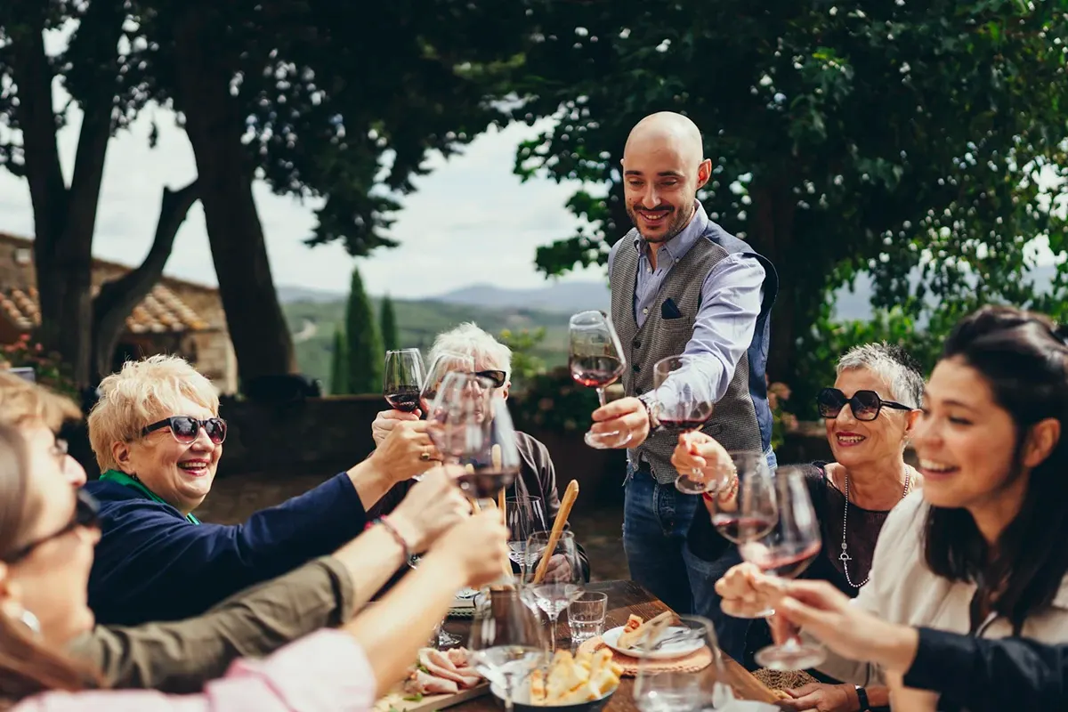 Guests raising glasses of red wine in a toast at an outdoor table under trees, with rolling hills behind