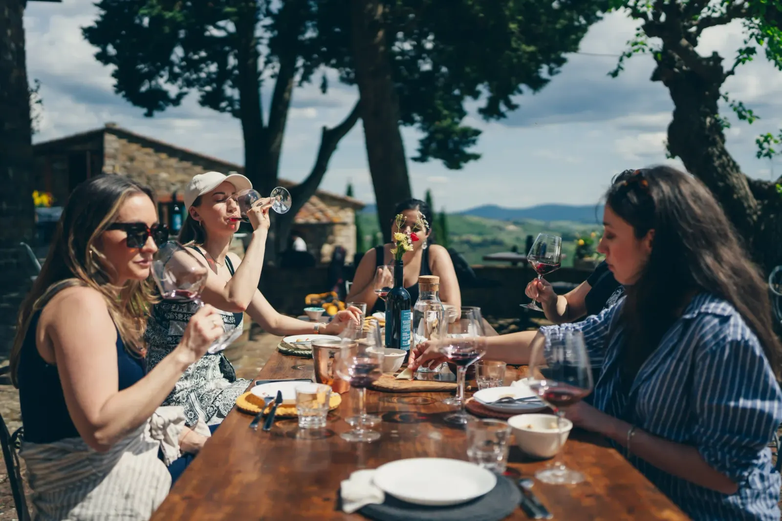 Women raising wine glasses at a long outdoor table set for lunch in the countryside