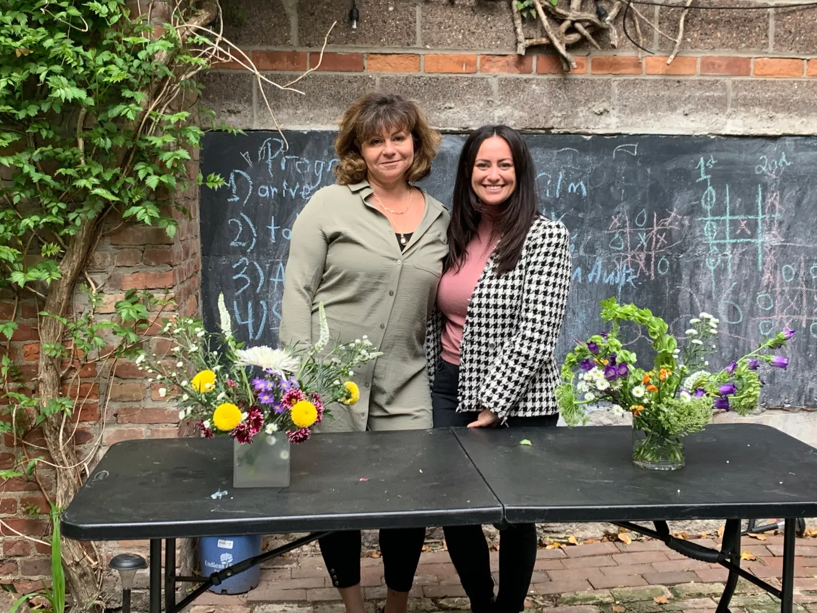 Two women posing behind a black table with two finished flower arrangements in glass vases in a brick-walled courtyard