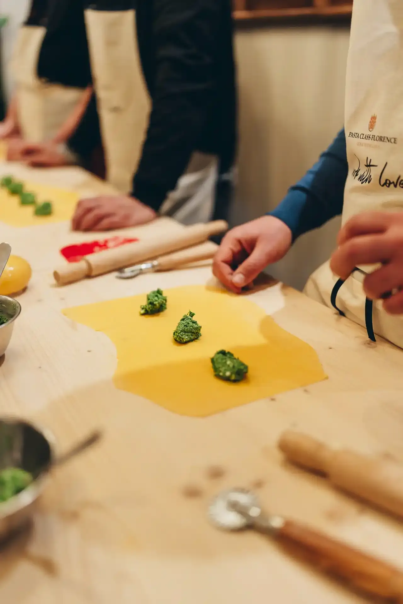 Close-up van handen die geel pastadeeg uitrollen met een deegroller, naast kleine hoopjes spinazievulling