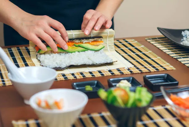 Hands rolling sushi on a bamboo mat, with bowls of fillings in front